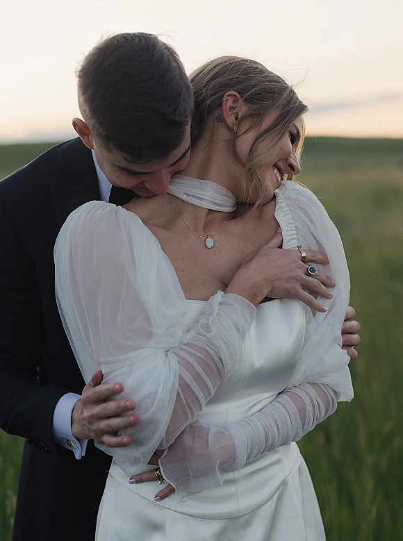 Couple portrait of bride and groom embrace as he hugs her from behind, her sheer-sleeve dress glowing in a sunset field