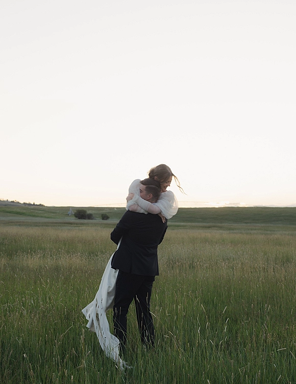 Couple portrait of bride and groom hug in an open meadow, her wedding veil trailing as he lifts her near rolling hills and horizon