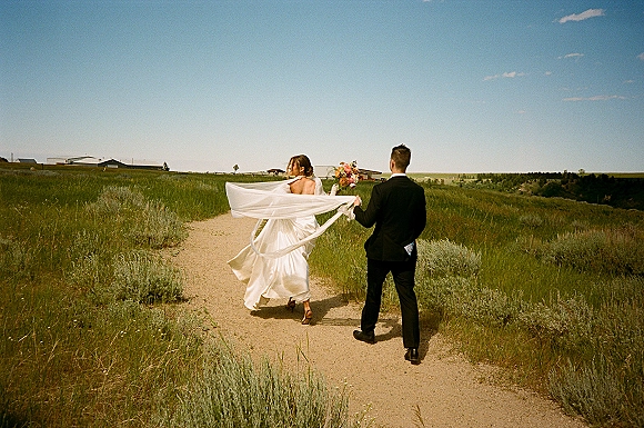 Couple portrait of bride and groom walking away on a dirt path, her veil blowing as she holds a bouquet under blue sky