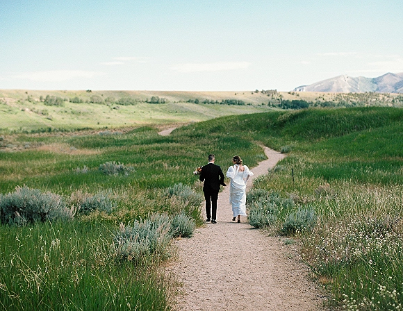 Couple walking away down a dirt path, bride and groom walking with bouquet and veil through a green meadow toward mountains under blue sky