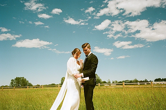 Couple portrait of bride in a flowing bridal cape and groom in a black tuxedo holding hands in a meadow by a wooden fence under blue sky