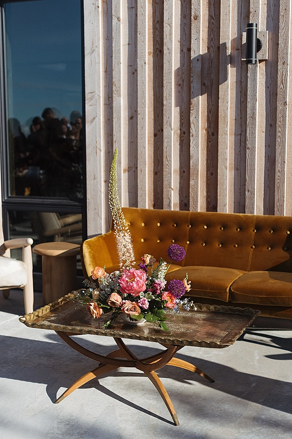 Wedding lounge decor with mustard velvet sofa beside a brass coffee table and floral centerpiece, set against a wood slat wall and window light