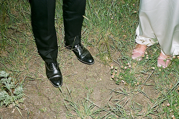 Wedding shoes detail with bride and groom shoes side by side, blush pink heels and black dress shoes beneath a white dress hem on clover grass