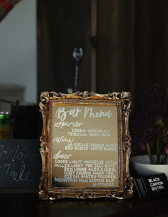 Wedding bar menu in an ornate gold frame with handwritten calligraphy beside flowers and black napkins at a dark bar counter