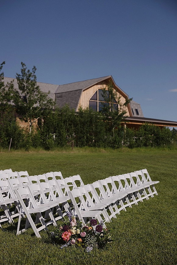 Ceremony seating with outdoor wedding chairs in neat rows of white folding chairs on a grassy lawn beside a rustic barn and trees under blue sky