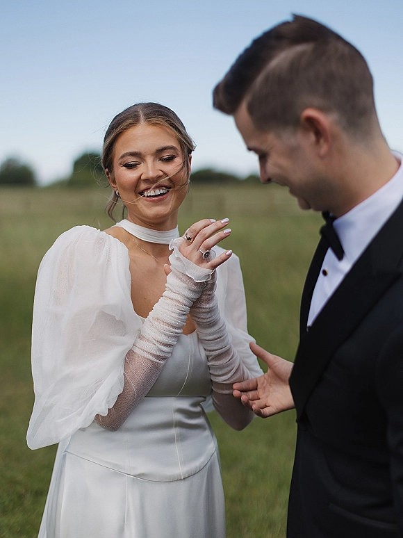 Couple portrait of a laughing bride and groom candidly holding hands, showing wedding rings in a grassy field under open sky