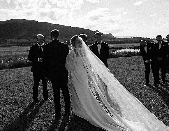 Ceremony moment as bride seen from behind in a long veil faces the altar, with tuxedoed wedding party by a lakeside mountain view