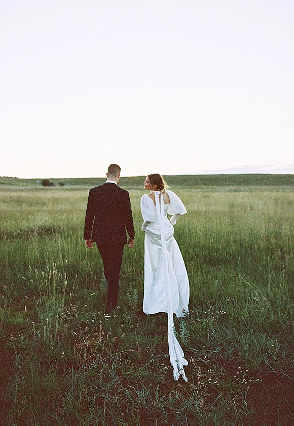 Couple portrait of bride and groom walking away in a meadow, bride glancing back as her long train trails across the grass