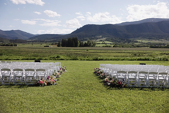 Ceremony setup for an outdoor wedding ceremony with white folding chairs and aisle florals on a meadow lawn with mountains beyond