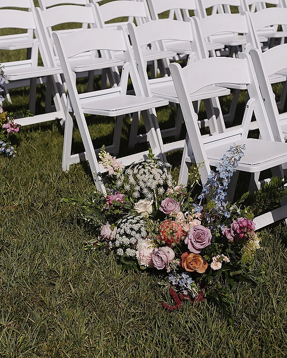 Ceremony chair decor with white folding chairs beside a ground floral arrangement of roses, blue delphinium, baby's breath and greenery on a lawn