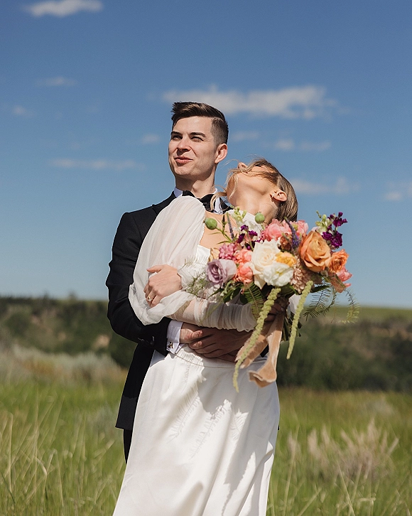 Couple portrait of bride and groom embrace, groom holding bride with a bright wedding bouquet in a grassy field under blue sky