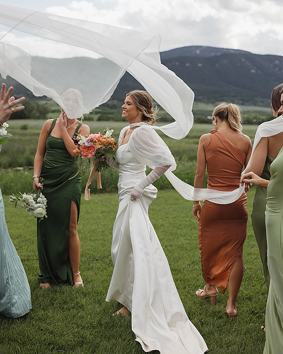 Bride with bridesmaids laughing as the bride holding bouquet walks in a grassy mountain meadow, veil blowing under a cloudy sky