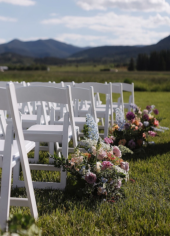 Ceremony seating with white folding chairs and aisle flowers on a grassy meadow, framed by mountain views, forest, and clouds above
