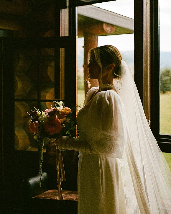 Bridal portrait of a bride holding bouquet in side profile by a window, veil and long-sleeve gown in a log cabin with mountain view