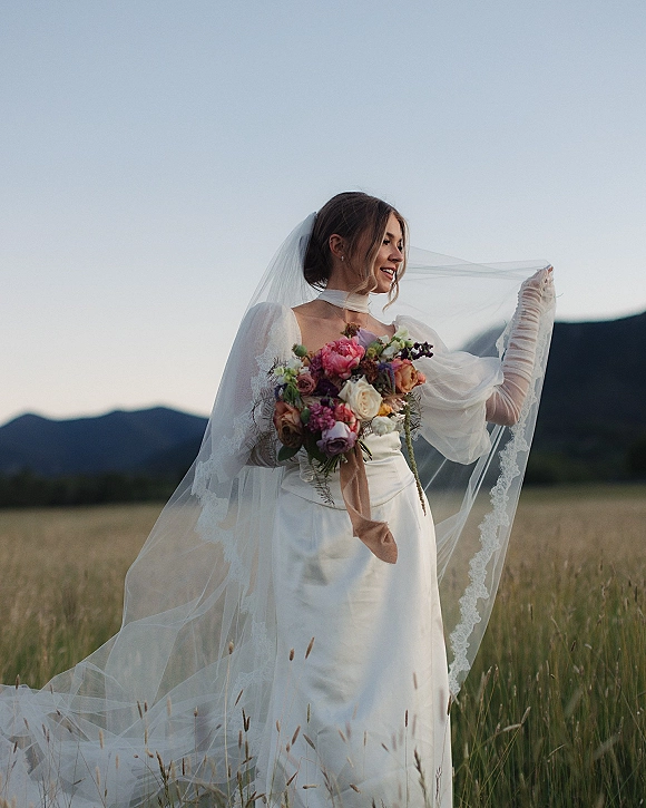 Bridal portrait of a bride holding bouquet, wearing a long lace-trim veil over her face in a meadow with mountains behind