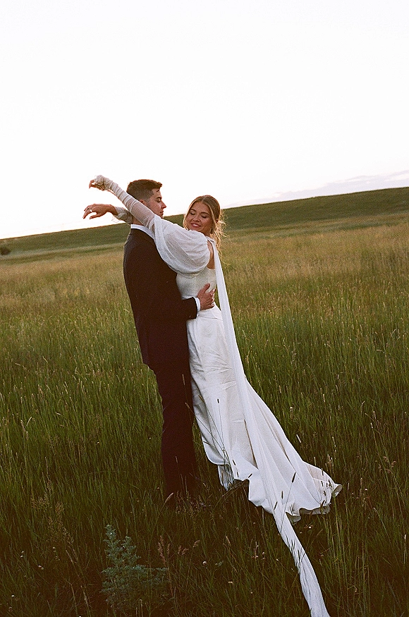 Couple portrait of bride in wedding dress and veil hugging groom in suit in tall grass field with rolling hills at sunset