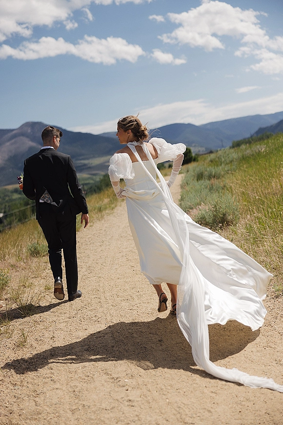Couple portrait of bride and groom walking away on a mountain path, her long train and veil blowing in the wind under blue skies