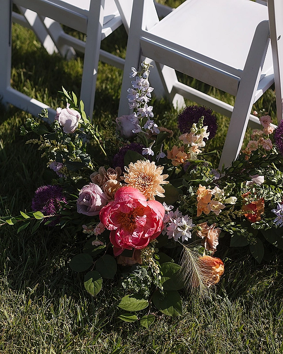 Aisle florals and ground aisle flowers with pastel peonies, roses, dahlias, and greenery beside white folding chairs on a sunlit lawn