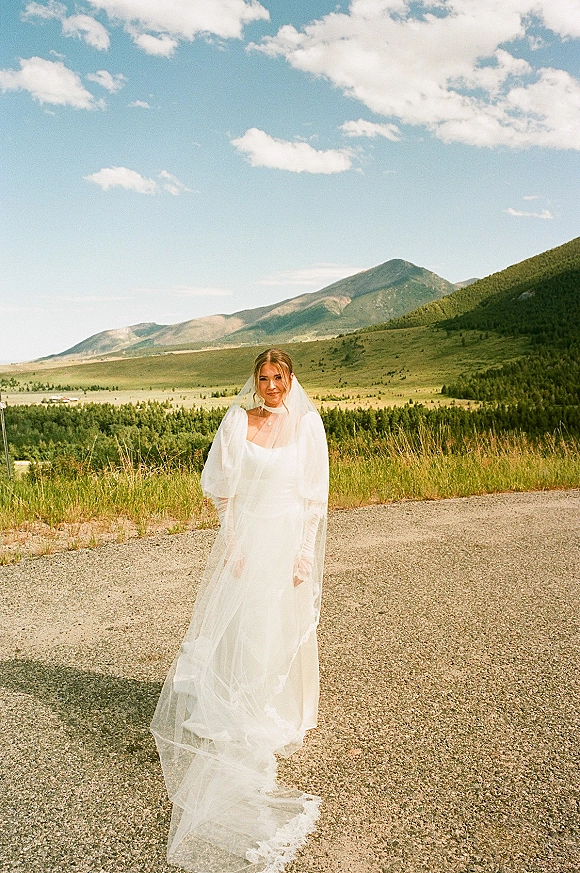 Bridal portrait of a bride in a veil on a gravel road, her tulle gown and train flowing with mountains, meadow, and pine trees behind