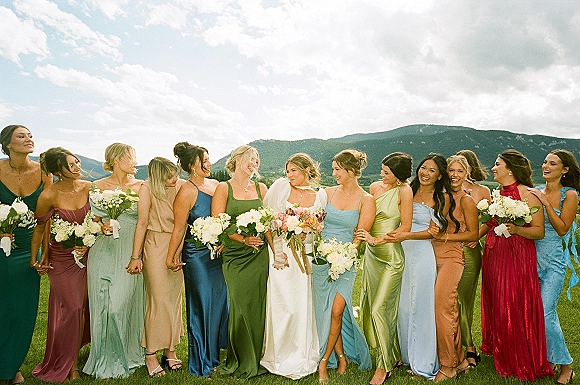 Bridesmaids group photo with bride in satin gown, holding bouquets in colorful dresses, standing in a grassy field with mountains behind
