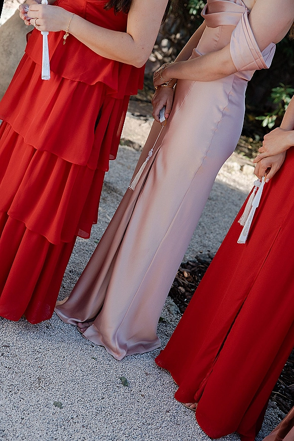 Bridesmaid dresses in mixed bridesmaid dress colors, including a champagne satin gown and ruffle styles, with tassel fan programs on a gravel path