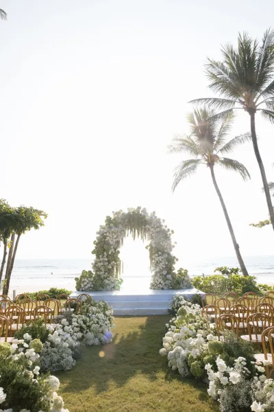 Ceremony setup for a beach wedding ceremony centered on a floral arch, overlooking the ocean with palm trees along the lawn.