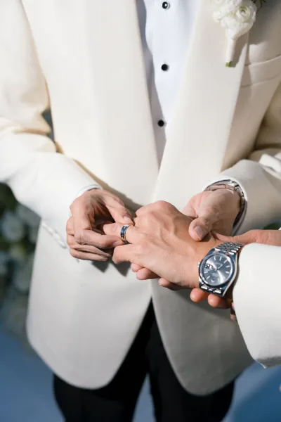 Ring exchange captured as a wedding ceremony detail, groom's hands trade simple wedding bands, accented by a white boutonniere.
