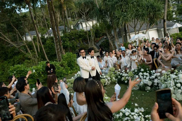 Ceremony recessional at a two grooms wedding, walking hand in hand through confetti as guests cheer amid palm trees in a tropical resort garden.