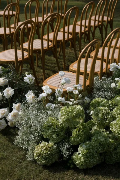 Ceremony aisle design for a garden ceremony setup, lined with bentwood chairs and bordered by white-and-green ground florals on a lawn.