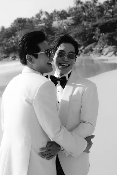 Black and white couple portrait of two grooms laughing and hugging in matching white tuxedos at a beach wedding, with palms and surf behind.