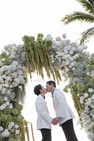 Wedding kiss portrait as two grooms kiss under a floral arch, framed by a sunlit palm tree and sky during a modern outdoor ceremony.