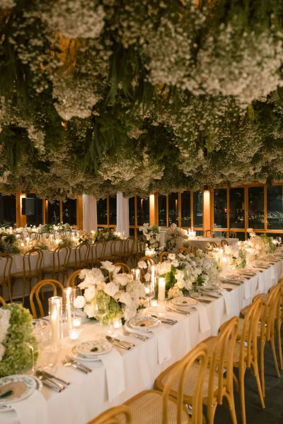 Reception tablescape showcasing long banquet table decor, with hanging florals overhead, set by wood-framed windows and drapery with a night view.