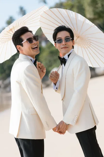 Couple portrait of two grooms holding hands on beach sand by the ocean, wearing sunglasses, laughing under a clear sky with trees beyond.