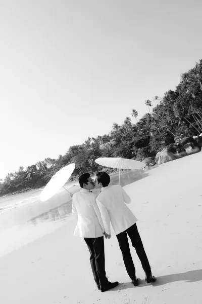 Wedding kiss portrait of a beach wedding couple, two grooms in white tuxedo jackets sharing a kiss by ocean waves and palm trees.