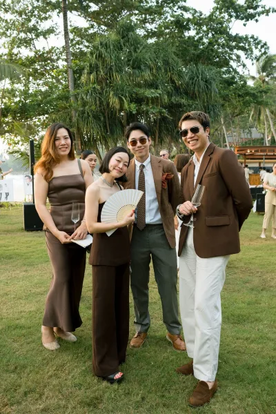 Wedding cocktail hour with outdoor reception guests mingling, raising champagne flutes by palm trees on a lawn near an outdoor bar.