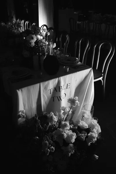 Reception tablescape, a candlelit reception table set in an indoor venue with wood floors and rows of chairs lining the background.