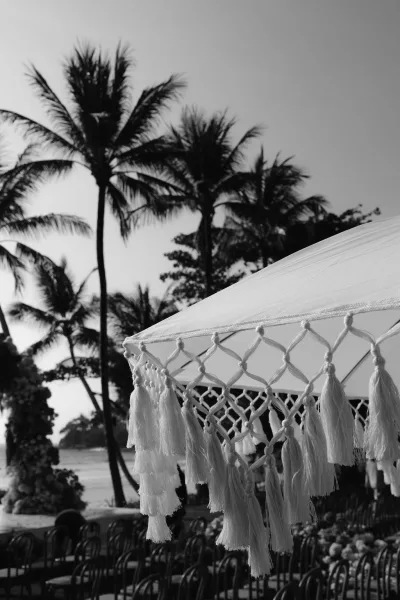 Beach ceremony decor anchors a tropical wedding ceremony with a macrame tassel umbrella, set against swaying palm trees and a gleaming ocean.