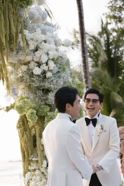 Ceremony moment as two grooms holding hands stand beneath a floral arch, laughing at the altar with palm trees and ocean behind.