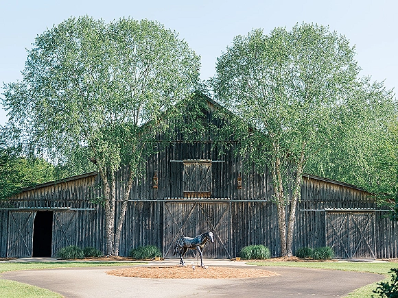 Barn wedding venue with rustic barn wedding venue wood doors, lanterns, and a horse statue by a driveway, framed by trees and lawn