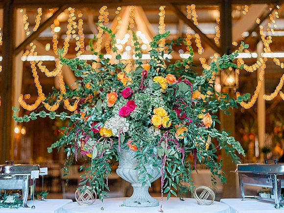 Wedding floral centerpiece in an urn floral centerpiece with roses and lush greenery on linen tables under string lights in a wood-beam hall