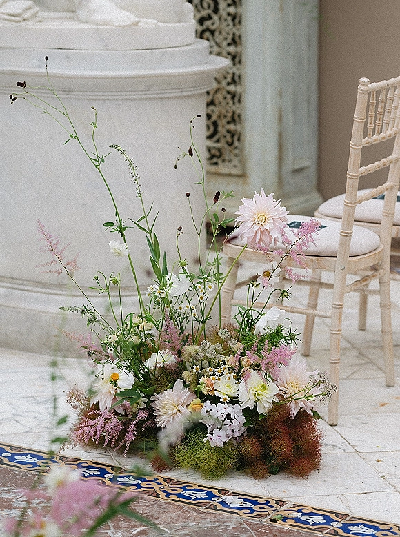 Ceremony aisle flowers in a ground floral arrangement with meadow blooms, dahlias and greenery beside a chiavari chair on tiled stone floor