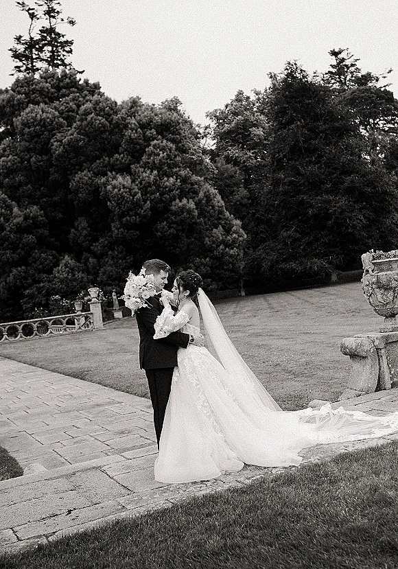 Couple portrait in black and white wedding portrait style, bride touching groom’s face, holding bouquet on a stone terrace garden lawn.