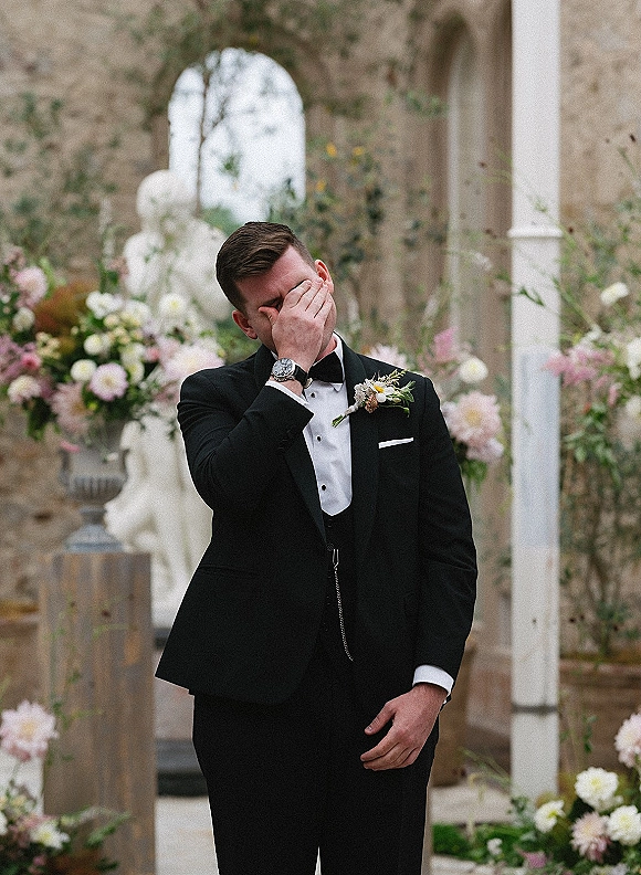 Groom portrait of an emotional groom wiping tears in a black tuxedo with boutonniere beside a stone church arched doorway altar