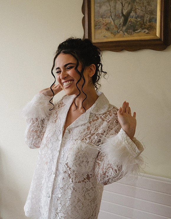 Bridal portrait of a getting ready bride in a lace robe, smiling with an updo and loose curls against a cream wall with wainscoting