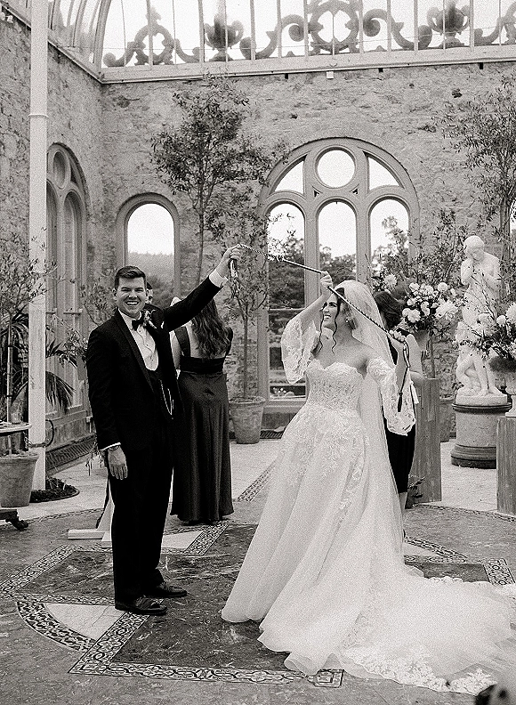 Wedding reception dance as bride twirls in lace veil while groom in tuxedo leads her in a stone courtyard with arched windows