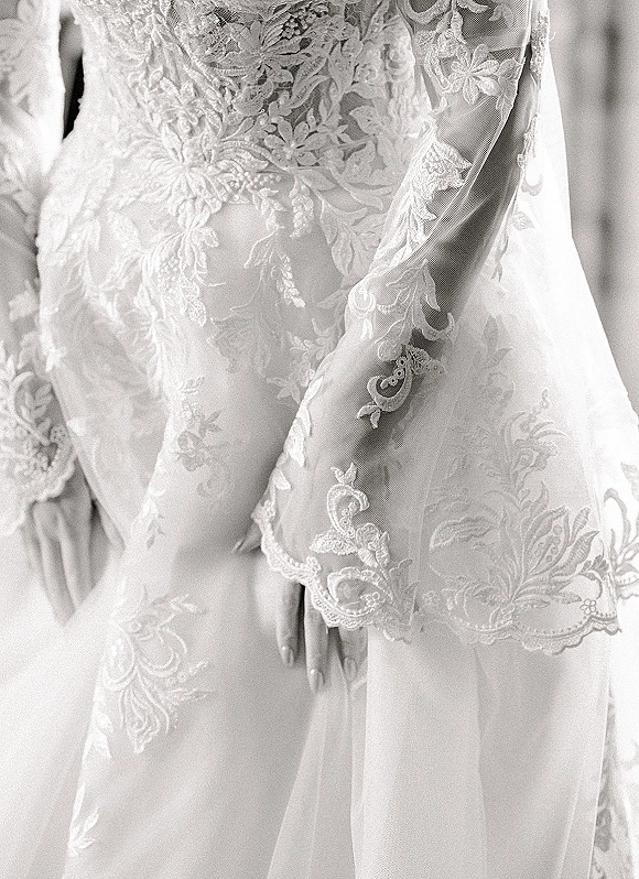 Wedding dress detail showing a lace wedding dress bodice with sheer illusion lace sleeves and floral embroidery against a neutral studio backdrop