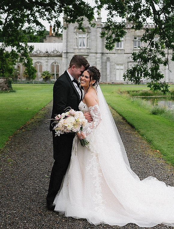 Couple portrait of bride and groom embrace with a forehead touch, lace off-shoulder gown and veil, holding bouquet on gravel path by a manor house