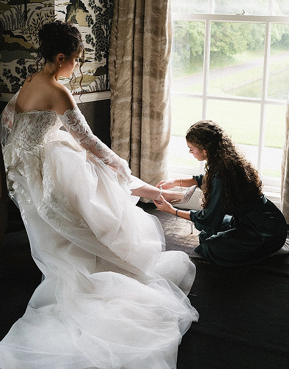 Bridal getting ready as a bridesmaid helps the bride put on heels by a window, off-the-shoulder lace dress glowing in soft light