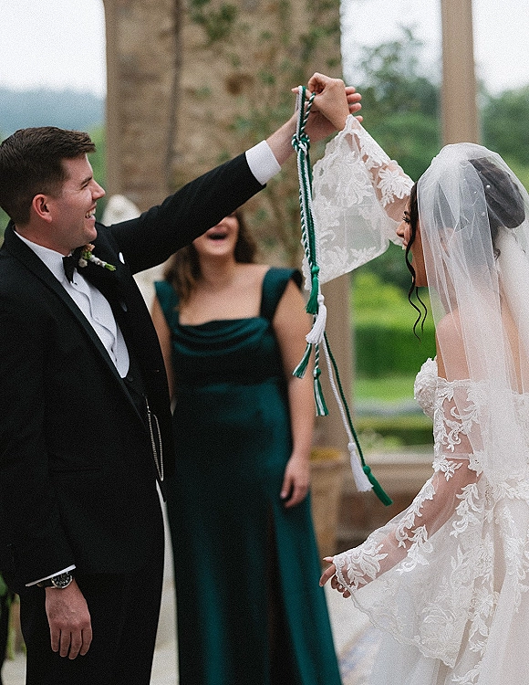 Ceremony moment during wedding handfasting as bride in lace dress and veil and groom in tuxedo tie corded hands by stone columns outdoors