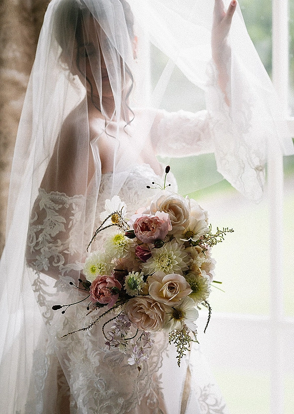 Bridal portrait of a bride holding bouquet of blush roses and dahlias, lace wedding dress and veil glowing in soft window light indoors